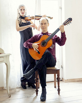 Ein Mann spielt Gitarre auf einem Stuhl, dahinter eine Frau, die Violine spielt