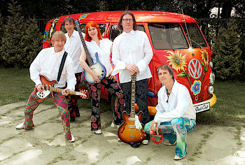 5 bunt gekleidete Musiker im Stil der 1960er- und 70er-Jahre vor einem roten VW-Bus