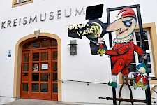 Kasperfigur vor einem alten Gebäude, auf dem KREISMUSEUM steht