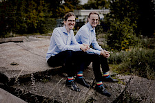 Zwei gleich mit blauem Hemd, schwarzer Hose und bunten Socken gekleidete schlanke Männer sitzen nebeneinander auf einer Art Ruine