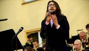 Junger Mann mit langen Haaren und Bart singt mit geschlossenen Augen in ein Mikrofon auf einer Bühne