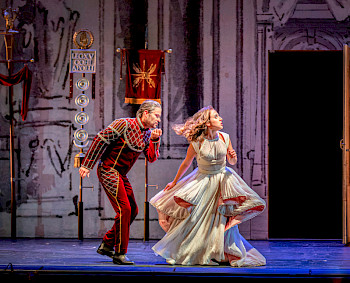 Eine Frau in weißem Tüllkeid neben einem Mann in rotem Fantasiekostüm auf einer Opernbühne