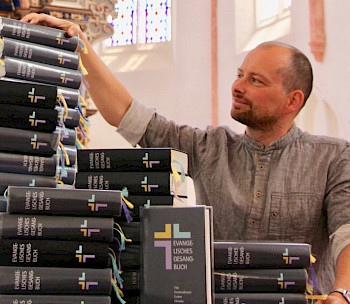 ein Mann greift glücklich auf einen Bücherstapel mit evangelischen Gesangbüchern