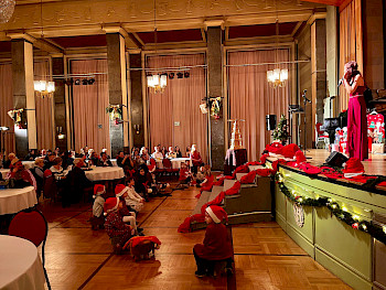 Eine Frau singt auf der Bühne. Die Kinder mit Klamotten von Weihnachtsmänner sitzen unter herum