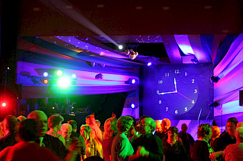 Ältere Paare tanzen auf Party in einem bunt beleuchteten Raum. Im Hintergrund ist eine große Uhr, die auf viertel vor zwölf steht.