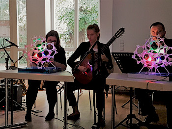 3 Musiker sitzen hinter Tischen. Eine spielt Gitarre, vor den anderen beiden steht eine bunt leuchtende, zellenartige Installation