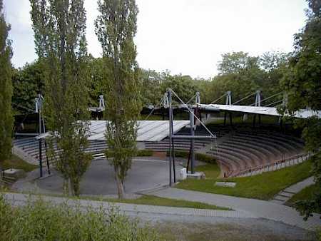 Ein Freiluft-Konzertort mit weißen Planen als Dächer, aus der Sicht auf den Zuschauerraum umgeben von Fichten