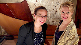 Eine brünette und eine blonde Frau stehen lächelnd vor einem Flügel