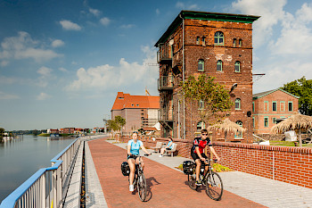Zwei Radfahrer in sommerlicher Kleidung fahren auf einer asphaltierten Promenade. Links ist ein Fluss, rechts ein historisches Backsteingebäude zu erkennen.