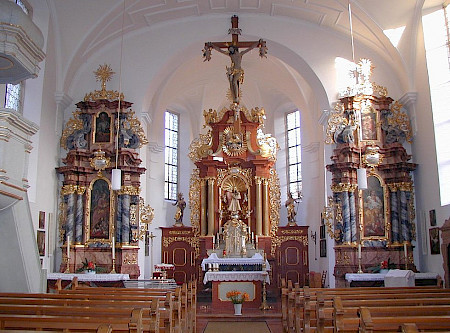 In einer Kirche, die weiße Wände hat, mit Blick auf einen Altar mit Kreuz, links und rechts braune Bänke, die leer sind