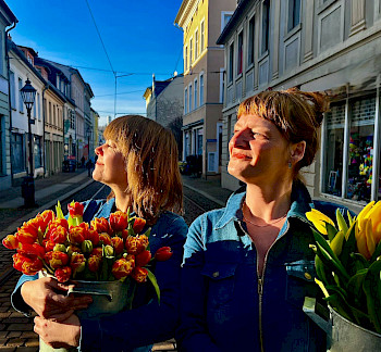 2 Frauen mit Blumen in den Armen, sie schauen in der Richtung von Sonnenscheinen
