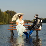 EIne Frau in weißem Kleid und weißen Schirm sitzt neben einem Taucher in Ausrüstung auf einer Sitzbank in einem See