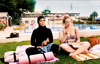 2 Frauen sitzen nebeneinander in der Schwimmhalle, 1 trägt Bikini, 1 bedeckt von islamischen Klamotten