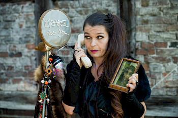 Eine Frau telefoniert vor einer Hauswand mit einem alten Kabeltelefon, in der Hand hat sie einen Bilderrahmen mit Foto