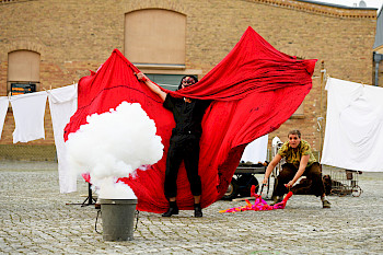 Mann wedelt mit einem großen rotem Tuch vor einer Wäscheleine, davor steht ein Topf, aus dem weißer Dampf steigt