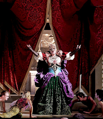 Frau in buntem, barocken Kostüm mit Maske breitet auf einer Bühne ihre Arme aus. Ihr zu Füßen liegen bäuchlings vier Männer und blicken zu ihr hinauf
