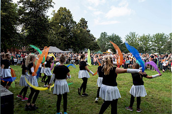 Eine Mädchentanzgruppe mit bunten Tüchern in den Händen tanzt auf einer Wiese vor Publikum