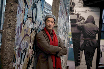 Mann mit Winterkleidung steht mit verschränkten Armen vor einer Lücke in der Berliner Mauer