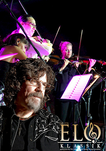 Hinter Phil Bates spielen die Musiker Geige in lilaer Licht