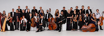 Ein Orchester sitzt vor dem weißen Hintergrund.