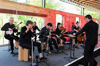 Das Ensemble Quillo ist auf dem Foto zu sehen. Die Musiker*innen spielen gerade auf ihren Instrumenten. Vorne rechts außen steht der Dirigent.