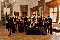 Etwa dreißig elegant schwarz gekleideter Musikerinnen und Musiker haben sich als Gruppe in einem schlossähnlichen Saal gruppiert