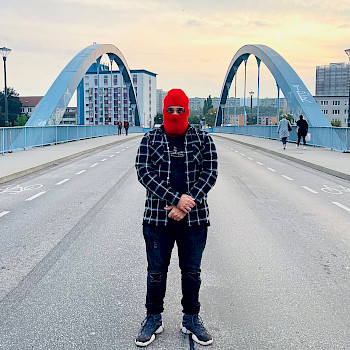 Ein schwarz gekleideter Mann mit einer roten Balaklava über dem Gesicht steht auf einer Autobrücke. Der Himmel ist bewölkt. Die Sonne geht unter. Man sieht ebenfalls Häuser im Hintergrund.