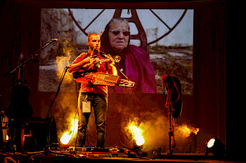 Omiri steht auf einer Bühne. Er hält in seiner linken Hand einen Geigenbogen. Um seinen Hals hängen zwei Instrumente. Im Hintergrund ist eine ältere Person auf einer Leinwand zu sehen.