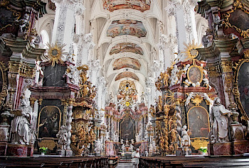 barocke Kirche von innen mit sehr vielen schnärkeligen Figuren, weiß und grau, mit Blick auf den Altar, davor eine weiß gekleidete Frau